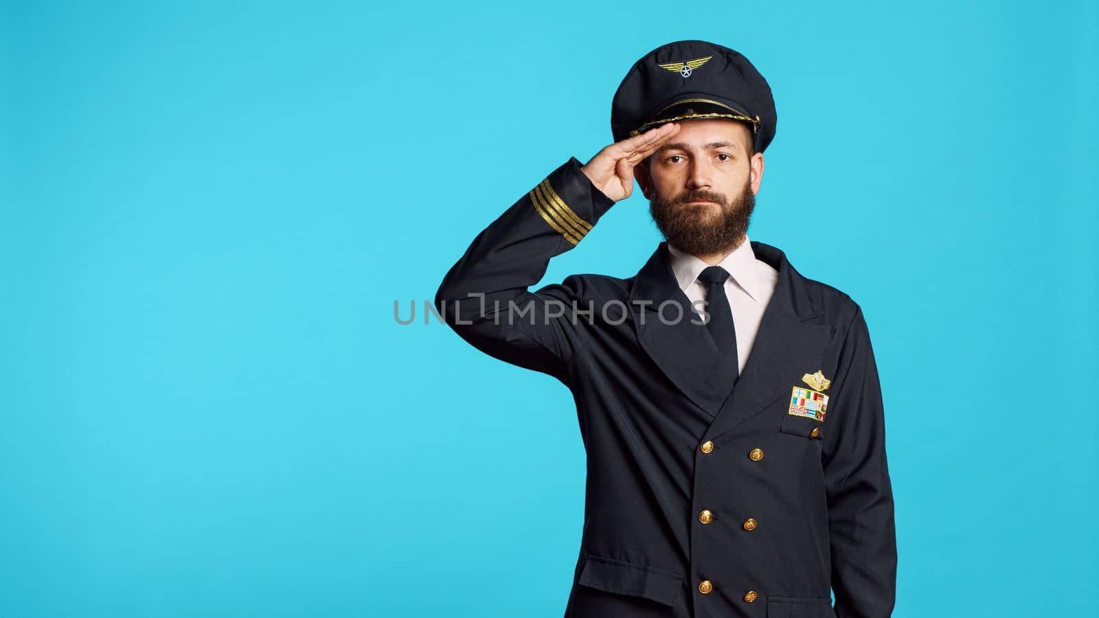 Male aviator doing military salute over blue backdrop, showing respect for army. Airplane captain serving flying industry as aviation worker, working on commercial flights and transportation.