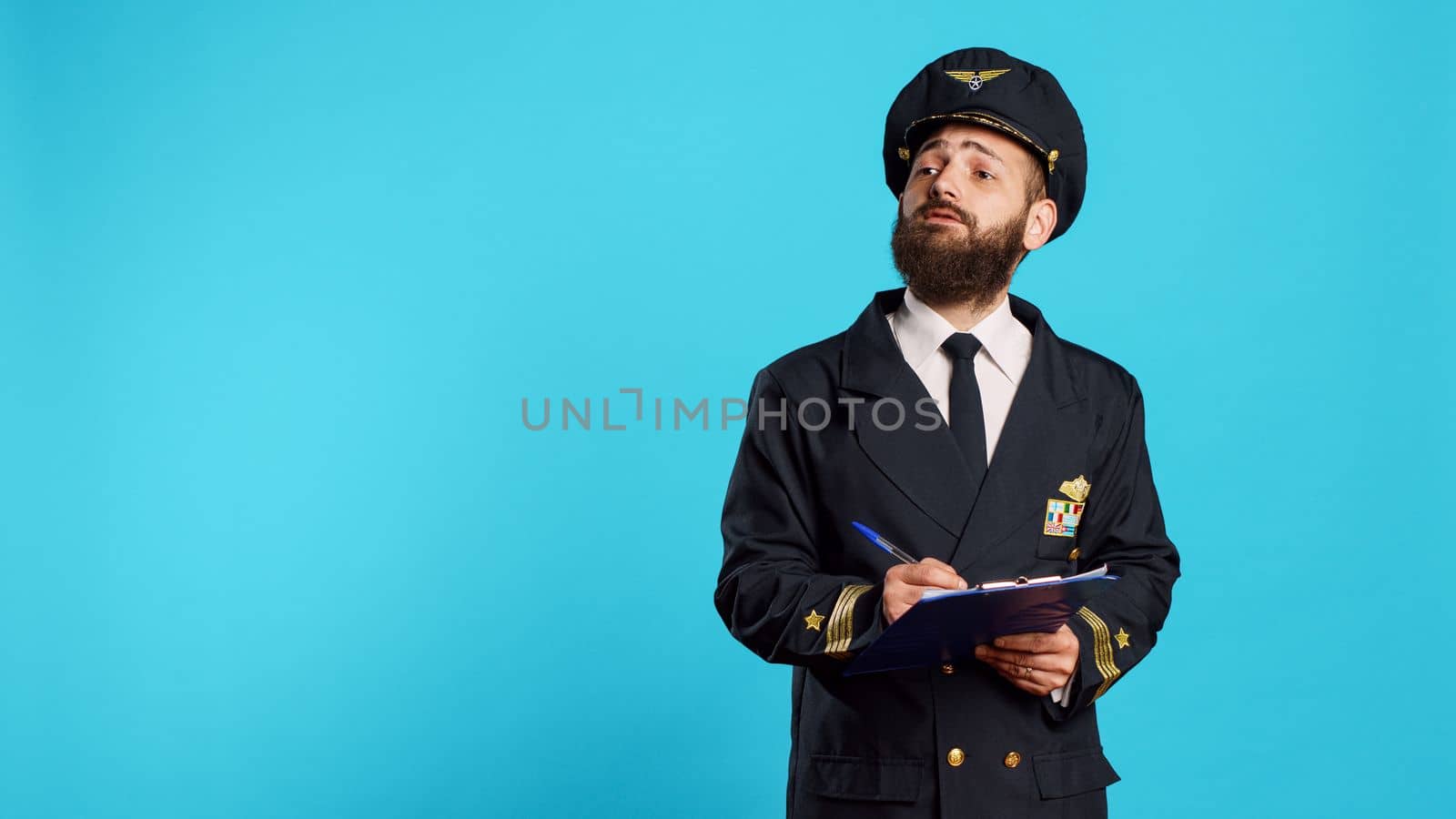 Confident aircrew captain checking list on papers, taking notes with pen and writing flying information. Man working as airplane pilot using clipboard files, wearing aviation uniform and hat.