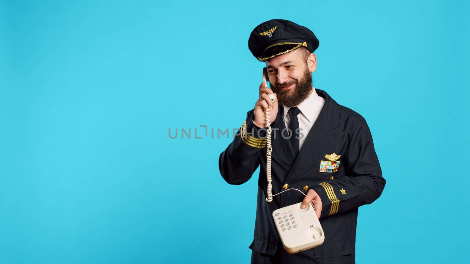 Commercial pilot talking on landline phone call in studio, answering office telephone with cord for remote conversation. Young smiling airliner in uniform chatting on phone line.