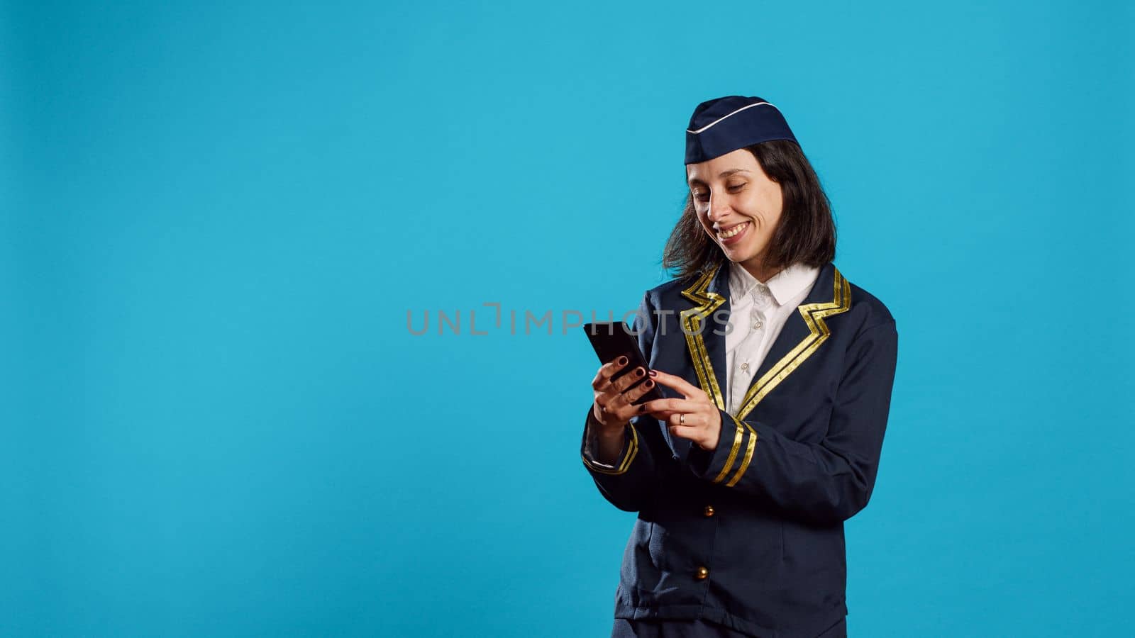 Aircrew member browsing online website on camera, using smartphone for social media app. Young air hostess in flying uniform playing with mobile phone and online browser over blue backdrop.