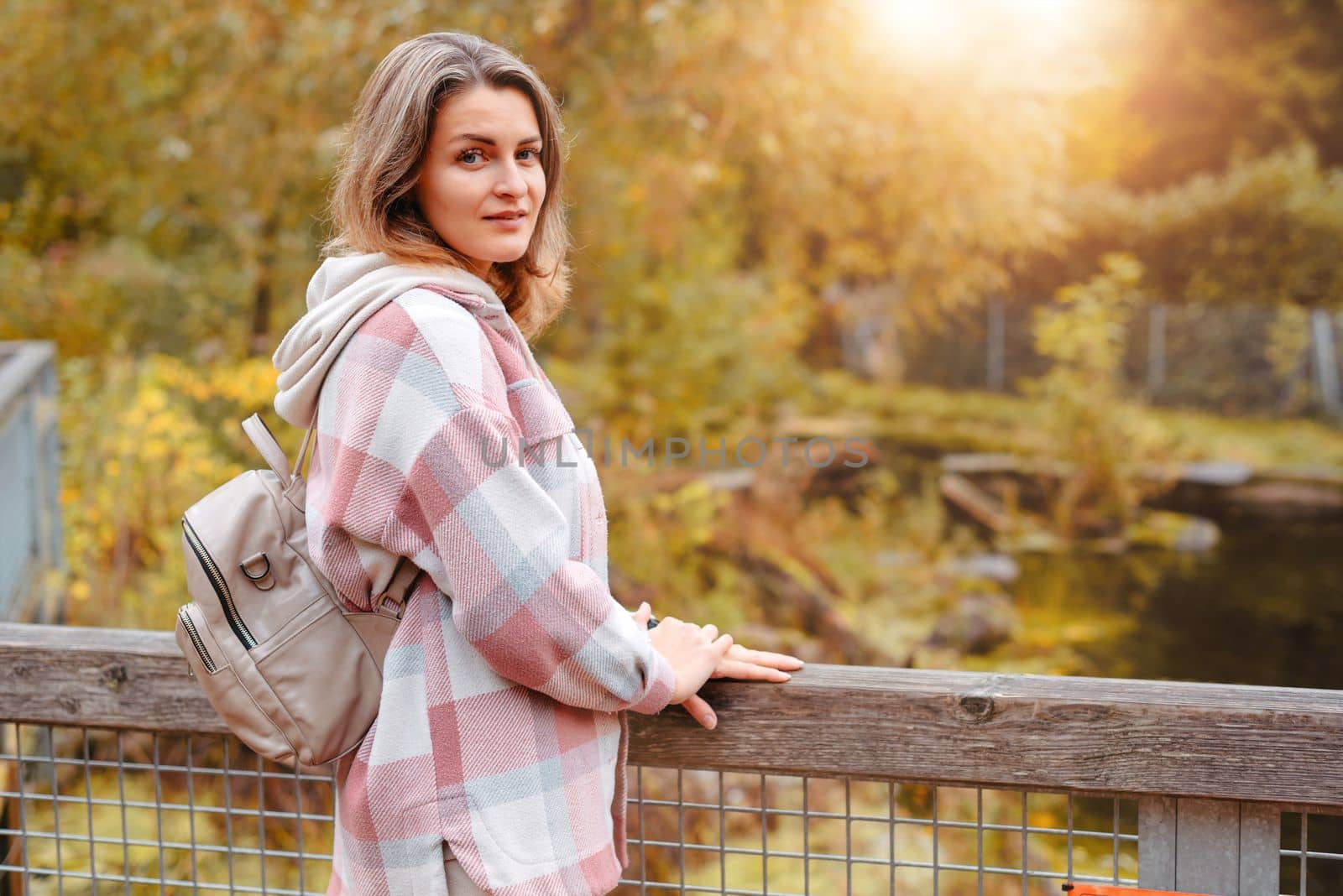 Portrait Of Cute Young Woman In Casual Wear In Autumn, Standing On Bridge Against Background Of An Autumn Park And River. Pretty Female Walking In Park In Golden Fall. Copy Space. Smiling Girl In The Park Standing On Wooden Bridge And Looking At The Camera In Autumn Season by Andrii_Ko