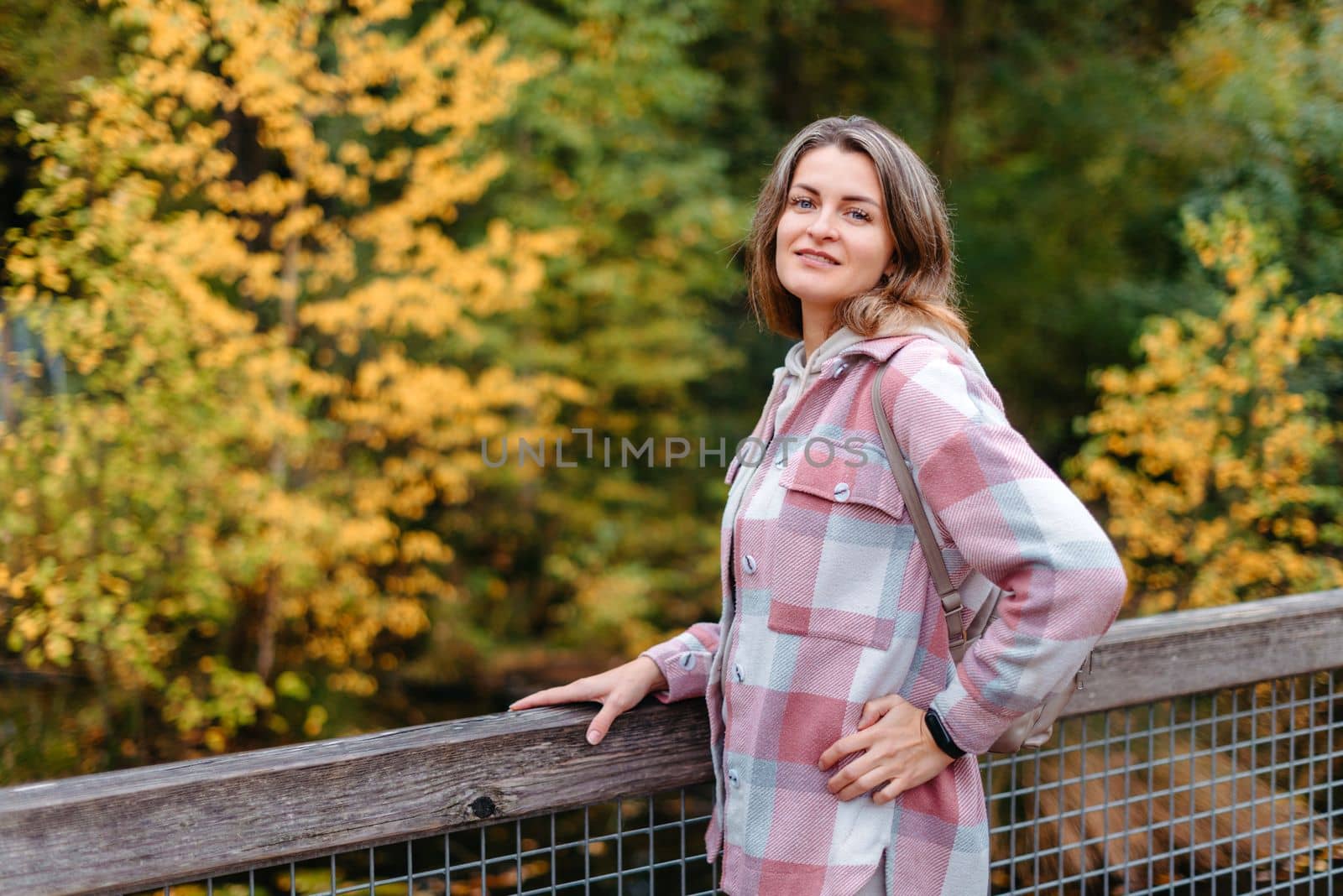 Portrait of cute young woman in casual wear in autumn, standing on bridge against background of an autumn Park and river. Pretty female walking in Park in golden fall. Copy space. smiling girl in the park standing on wooden bridge and looking at the camera in autumn season