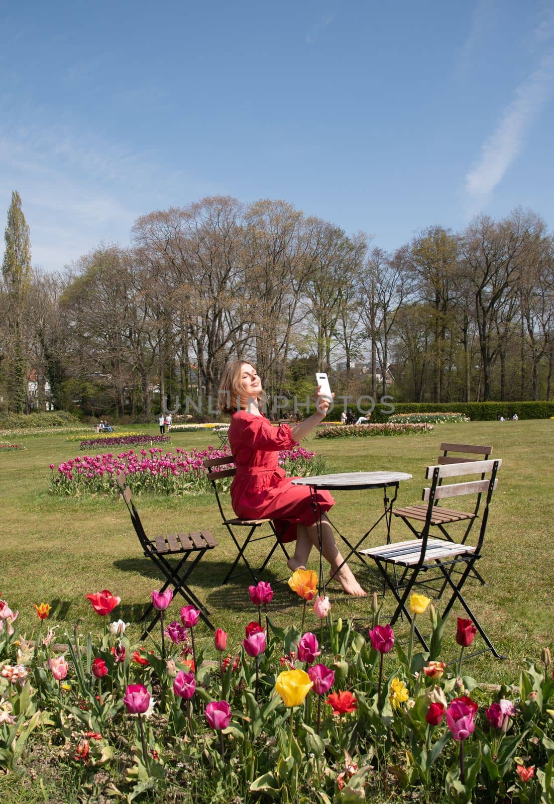 A young beautiful girl in a red dress in a spring park plays with her hair, flirts and takes a selfie on a mobile phone, tulips bloom and people walk in the park, pamper themselves.High quality photo