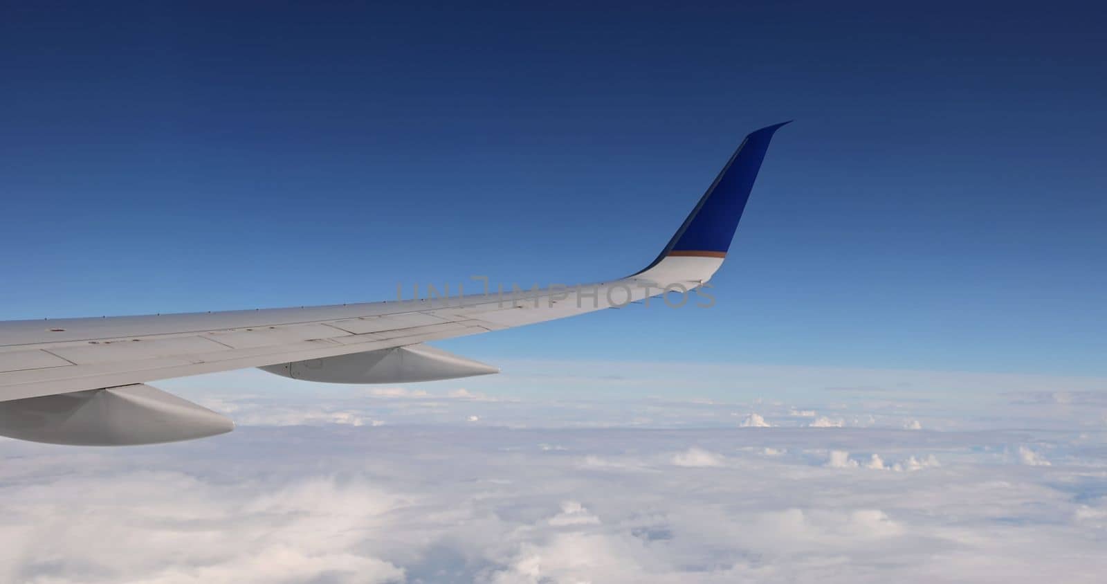 In background of fluffy cloudy sky, an airplane wing is seen flying above clouds skies from window