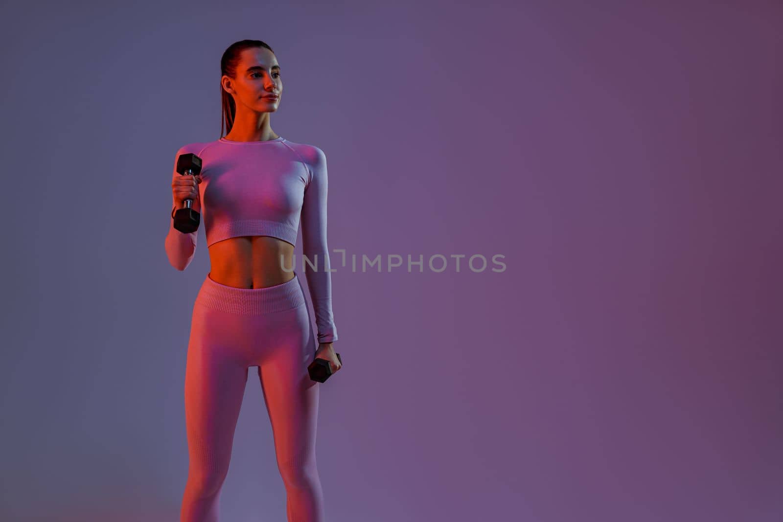 Attractive fitness woman doing exercises with dumbbells on studio background and looking at side by Yaroslav_astakhov