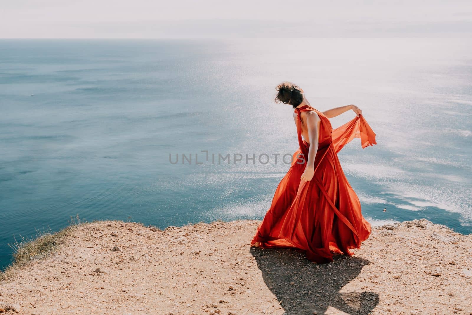 Woman in red dress on sea. Side view a Young beautiful sensual woman in a red long dress posing on a rock high above the sea on sunset. Girl on the nature on blue sky background. Fashion photo. by panophotograph