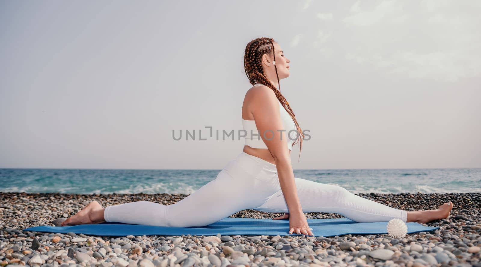 Woman yoga sea. Well looking middle aged woman with braids dreadlocks in white leggings and tops doing stretching pilates on yoga mat near sea. Female fitness yoga routine concept. Healthy lifestyle