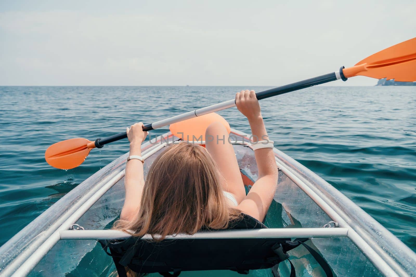 Woman in kayak back view. Happy young woman with long hair floating in transparent kayak on the crystal clear sea. Summer holiday vacation and cheerful female people having fun on the boat.