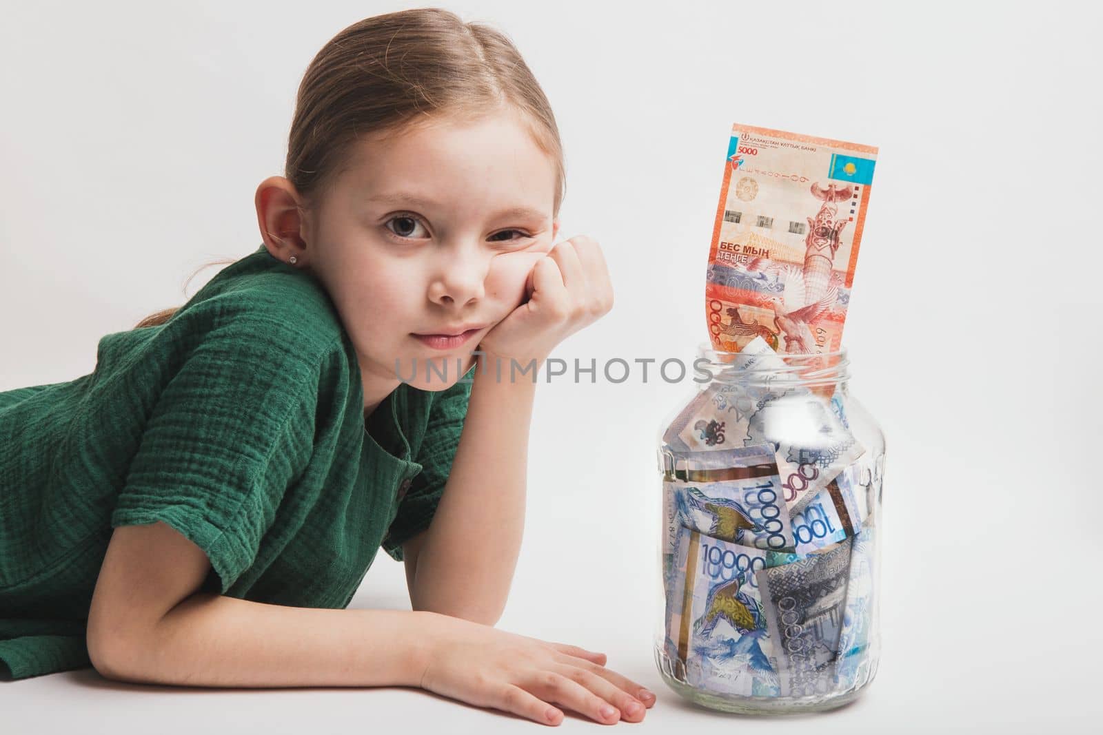 Sad and bored child with a large transparent jar with Kazakhstani tenge money on a white background.