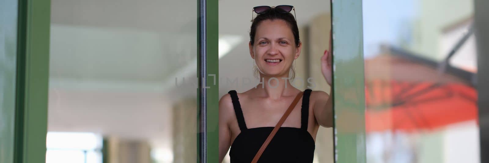 Beautiful smiling young woman opens glass door. Satisfied customer or meeting guests concept