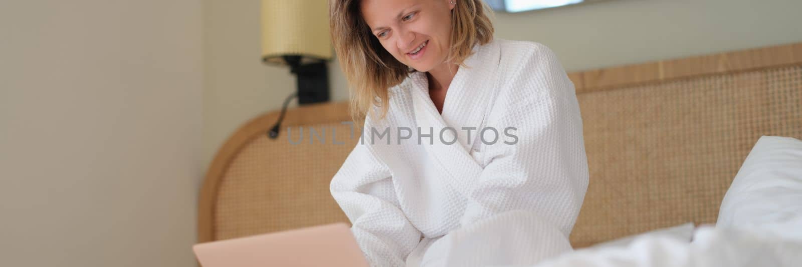 Woman sits at home on bed in white bathrobe and works on laptop. Work from home concept of quarantine and remote work freelancing