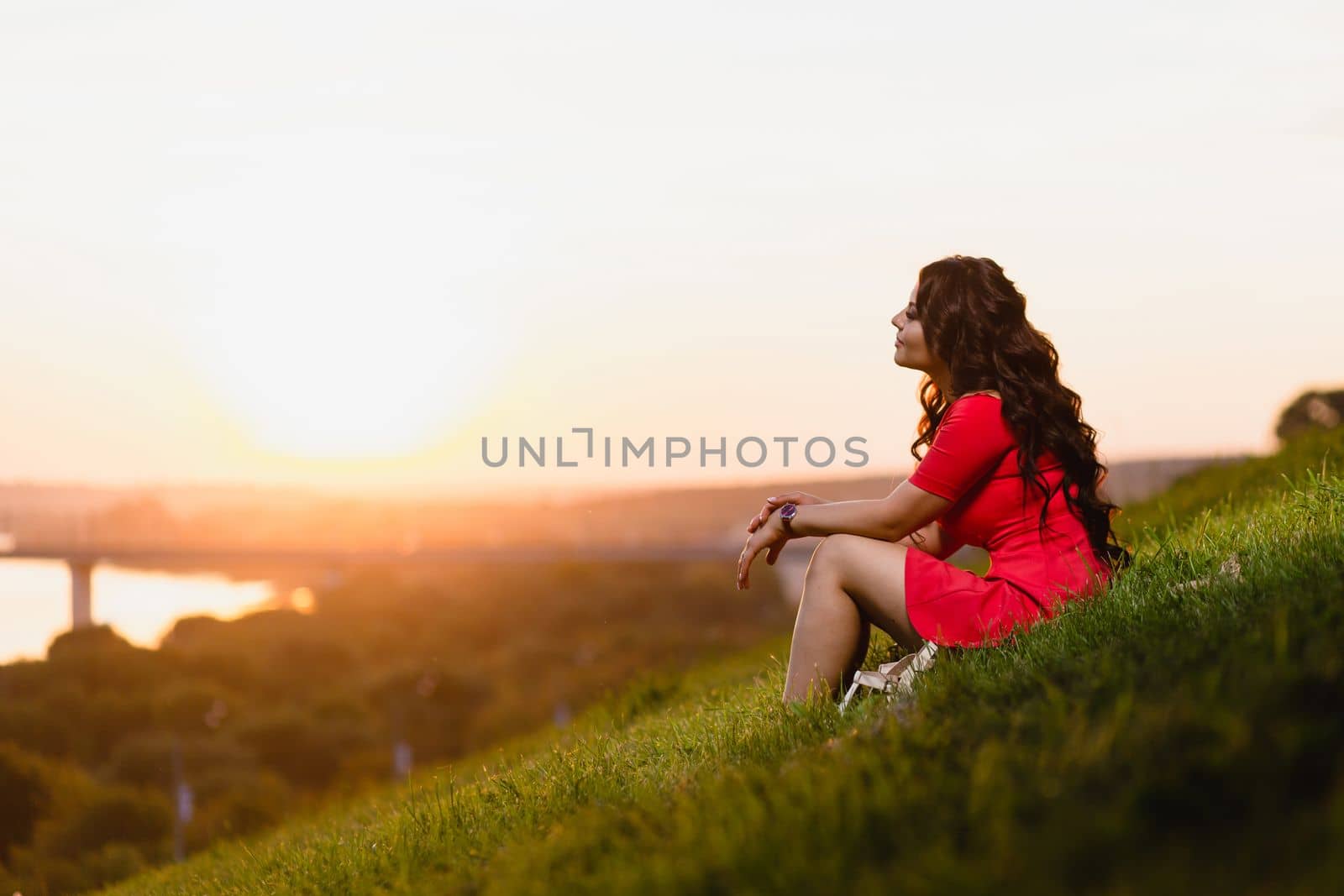 Beautiful young girl with long dark wavy hair sitting on a slope covered with green grass against the background of a summer sunset