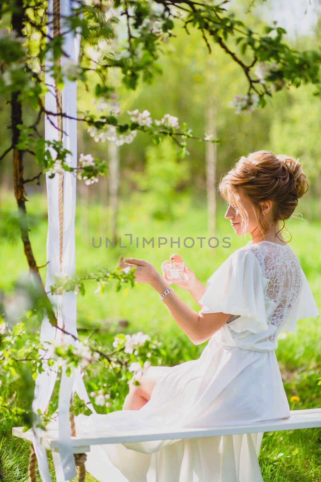 Young bride with blond hair in white negligee using perfume on a rope swing by Gennadii_Chebeliaev