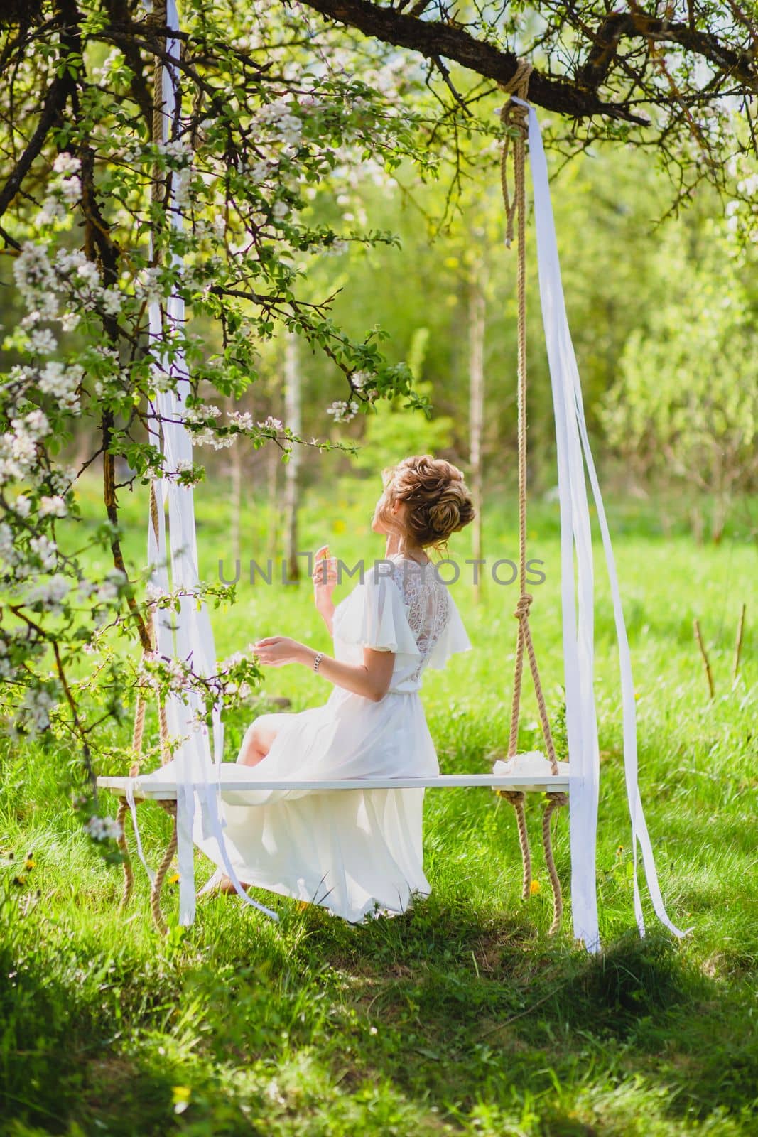 Young bride with blond hair in white negligee using perfume on a rope swing at the background of spring blossom orchard