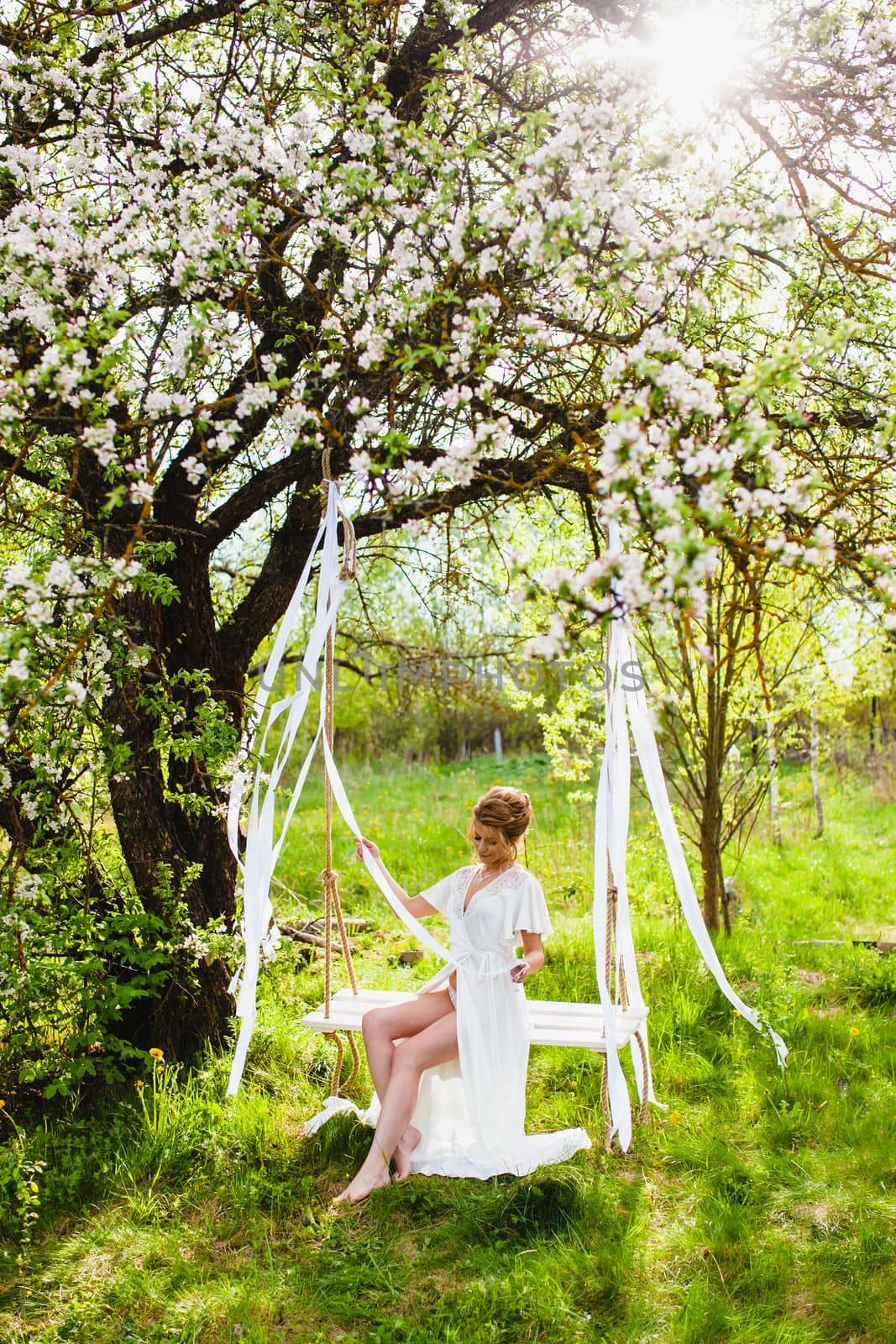 Young bride with blond hair and naked legs in white negligee sitting on a rope swing at the background of spring blossom orchard