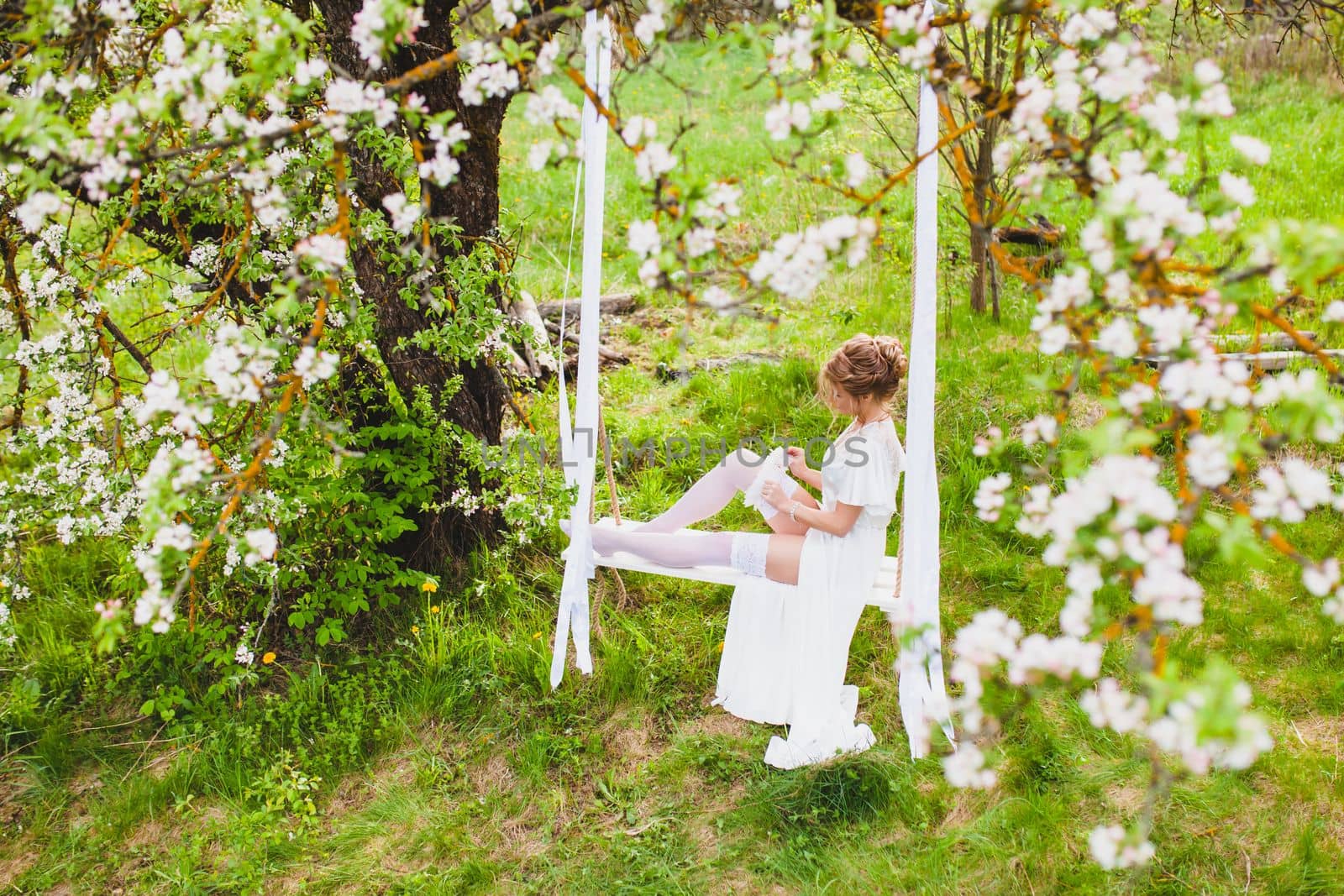 Young bride with blond hair in white negligee and stockings sitting on a rope swing putting on a garter at the background of spring blossom orchard