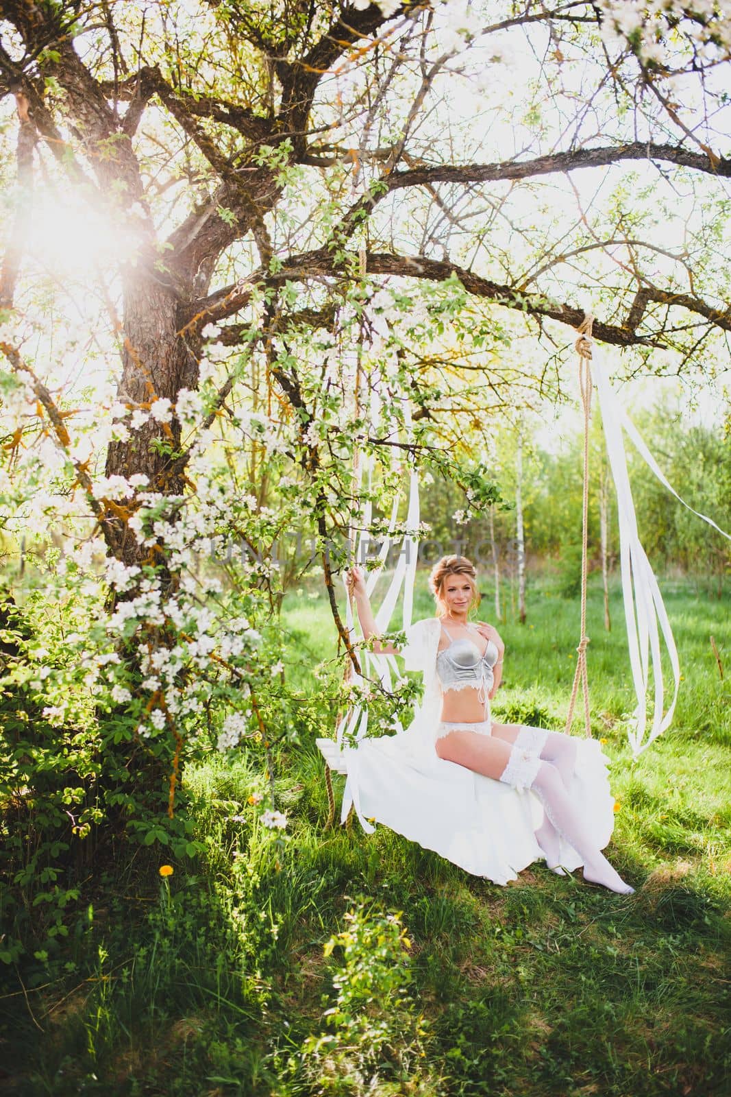 Young bride with blond hair in white negligee and stockings posing on a rope swing by Gennadii_Chebeliaev