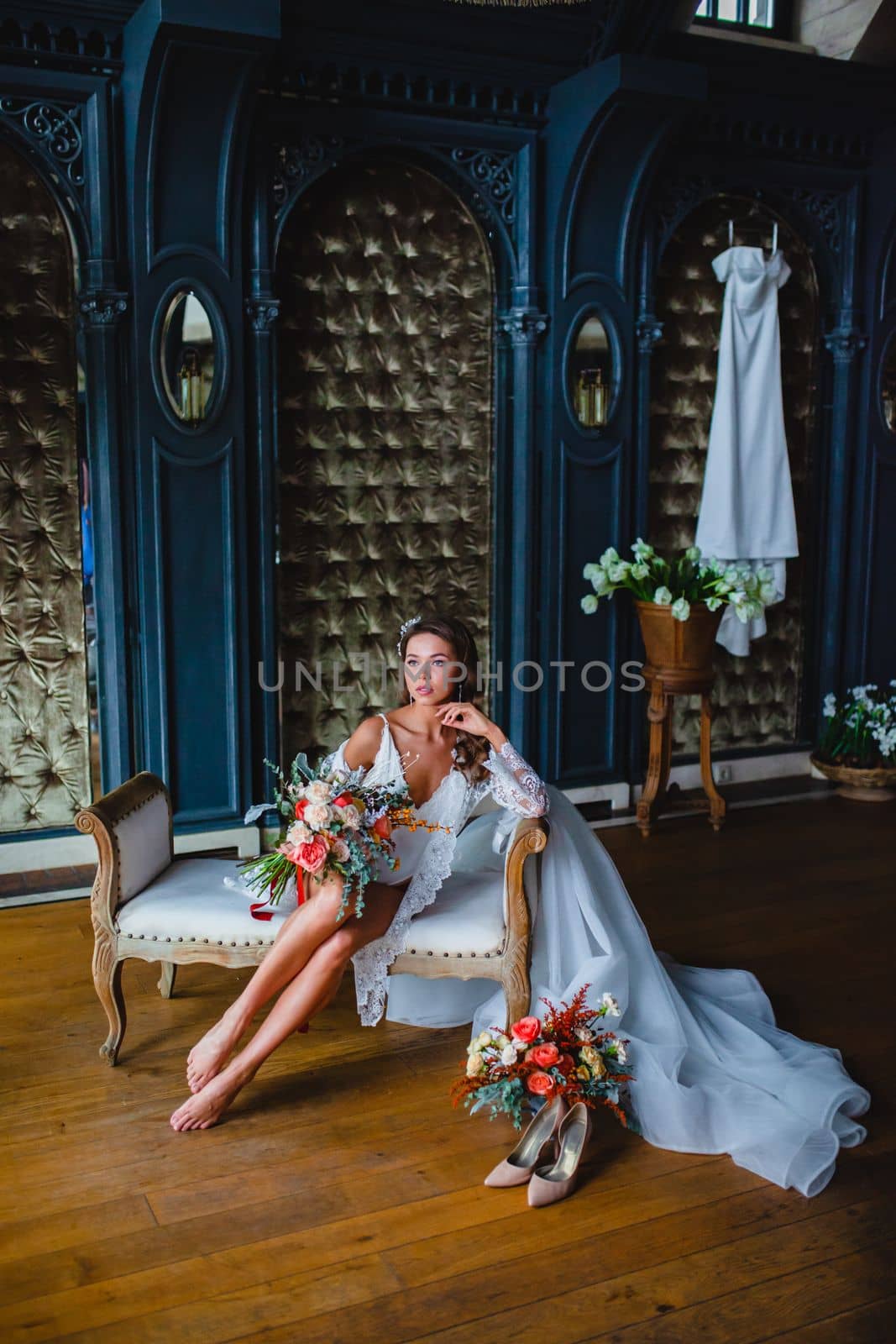 Seductive young bride with beautiful naked legs in white negligee and lingerie posing with gorgeous bridal bouquet on a sofa