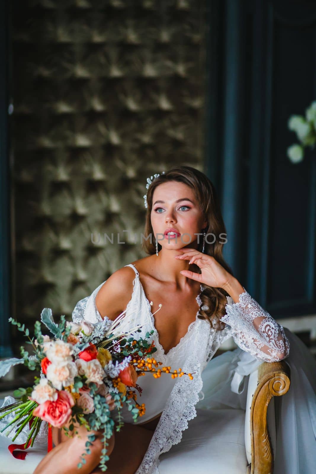 Close-up portrait of young bride in white negligee and lingerie posing with gorgeous bridal bouquet in the hotel room
