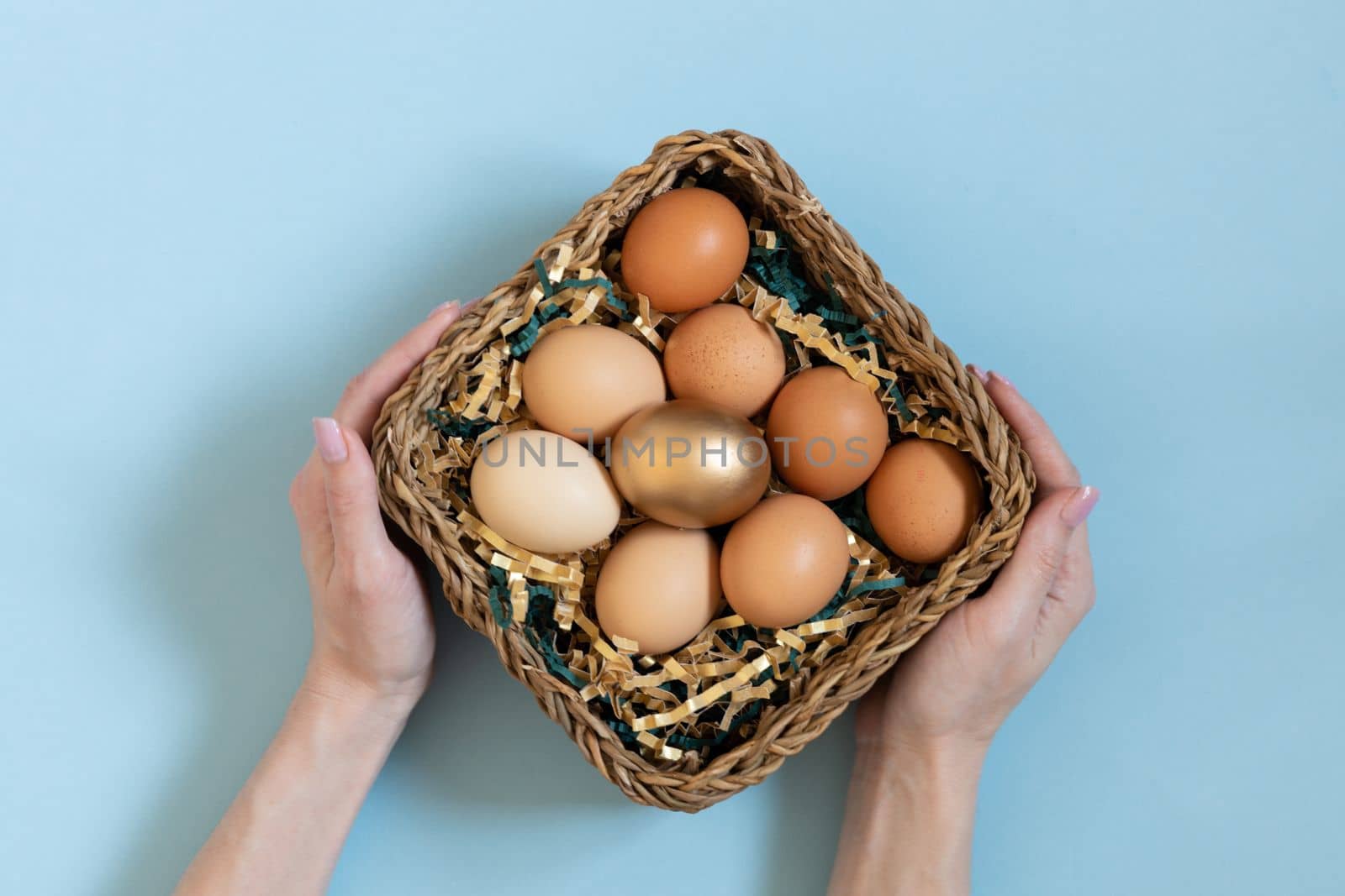 Gift basket with easter eggs. Women hands holding golden and natural color eggsin basket. Top view on pastel blue background. by Ri6ka