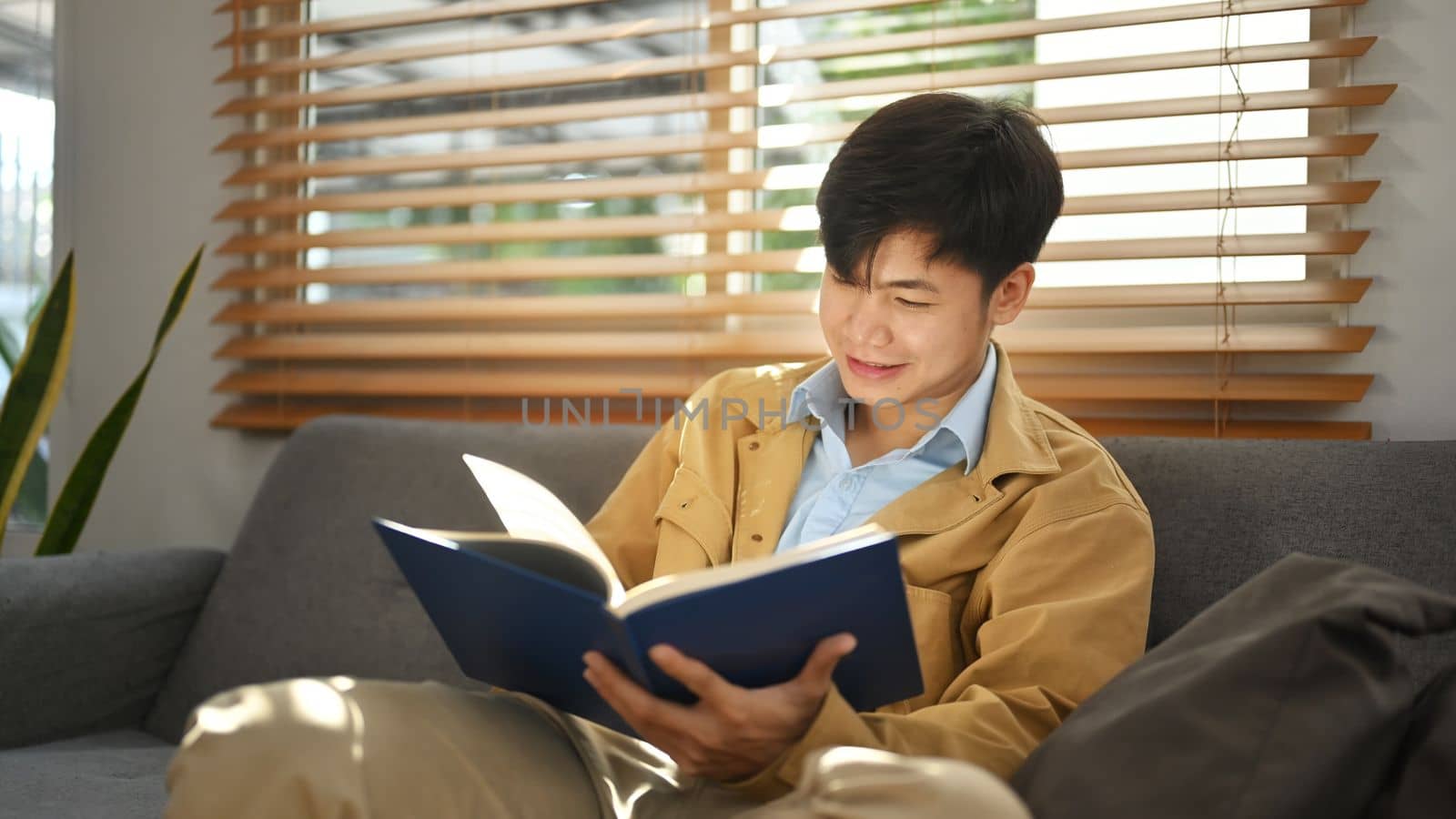 Satisfied man reading book, relaxing on comfortable couch at home. Leisure activity, positive mood concept by prathanchorruangsak