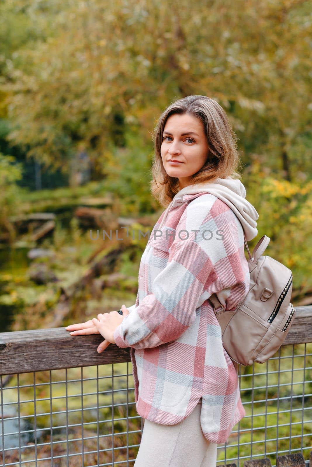 Portrait of cute young woman in casual wear in autumn, standing on bridge against background of an autumn Park and river. Pretty female walking in Park in golden fall. Copy space. smiling girl in the park standing on wooden bridge and looking at the camera in autumn season