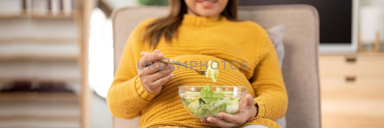 Happy pregnant young woman sitting and eating salad at home.