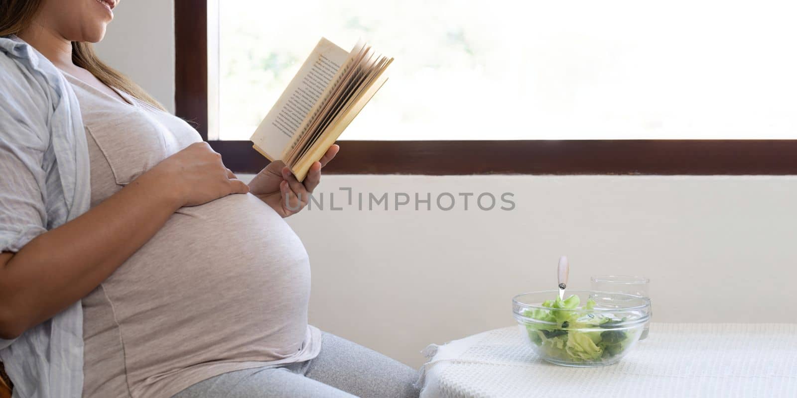 Young pregnant woman sitting reading a book and eating salad at home.