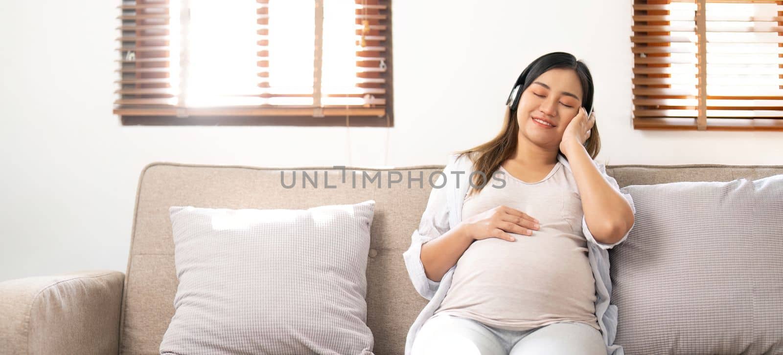 Happy Asian pregnant woman listening to music through her headphones on sofa. pregnancy lifestyle by nateemee