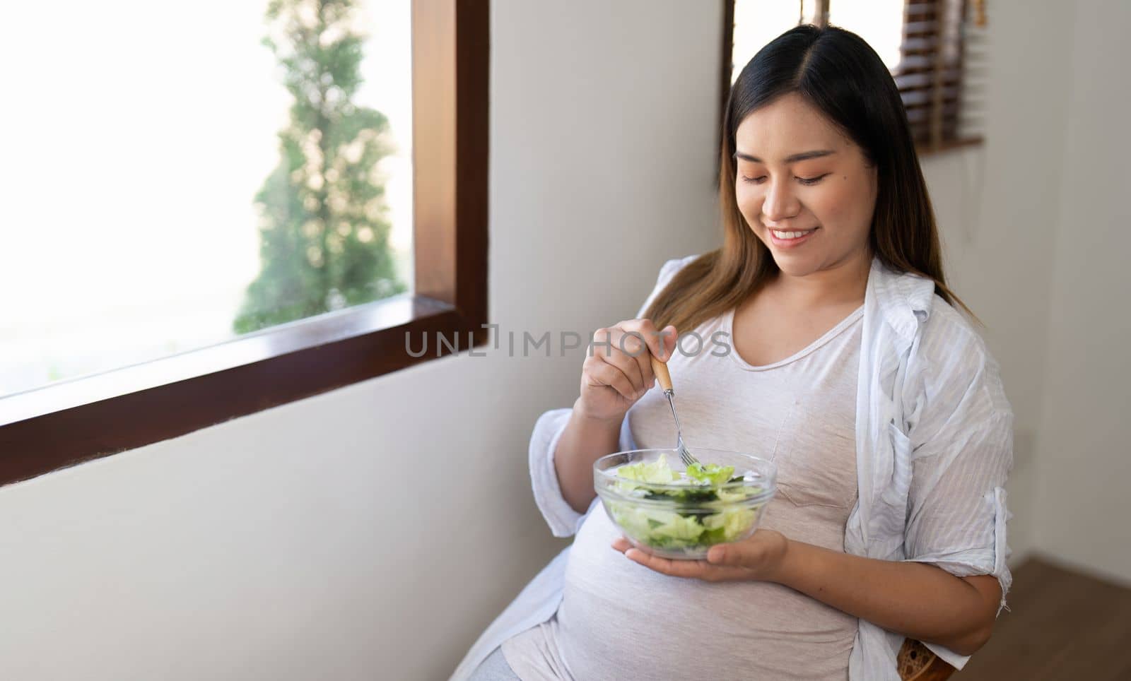 Happy pregnant young woman sitting and eating salad at home.