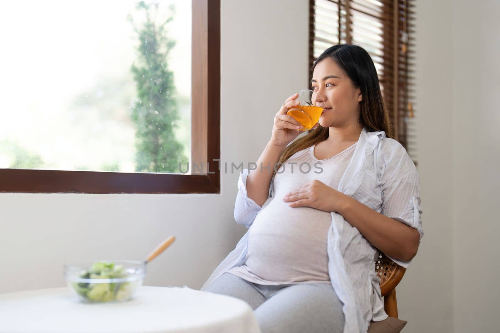 Happy pregnant young woman sitting and eating salad and orange juice at home by nateemee