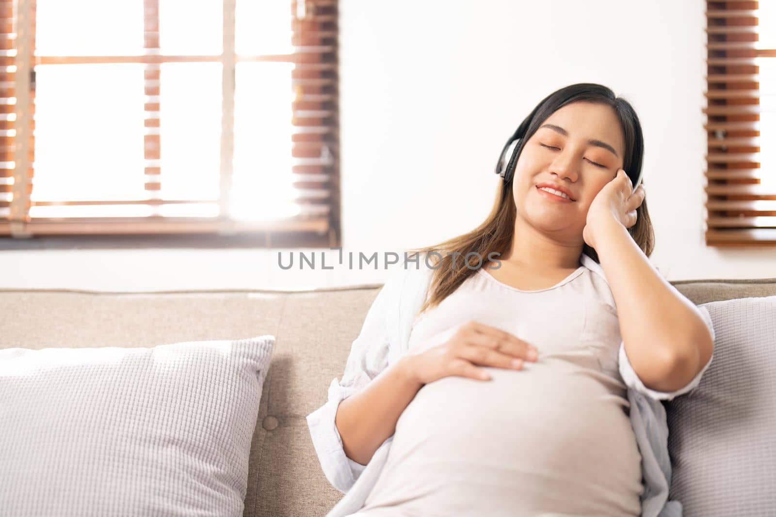 Happy Asian pregnant woman listening to music through her headphones on sofa. pregnancy lifestyle.