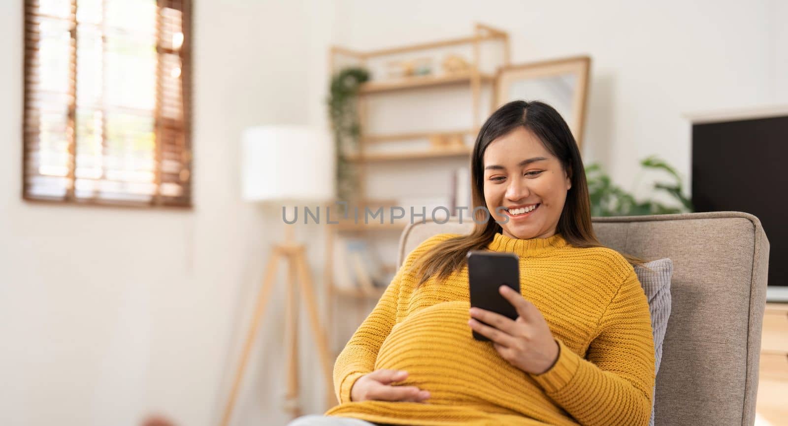 Happy Asian pregnant woman using her phone while relaxing on sofa in her living room by nateemee