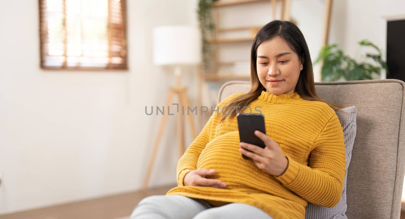 Happy Asian pregnant woman using her phone while relaxing on sofa in her living room.