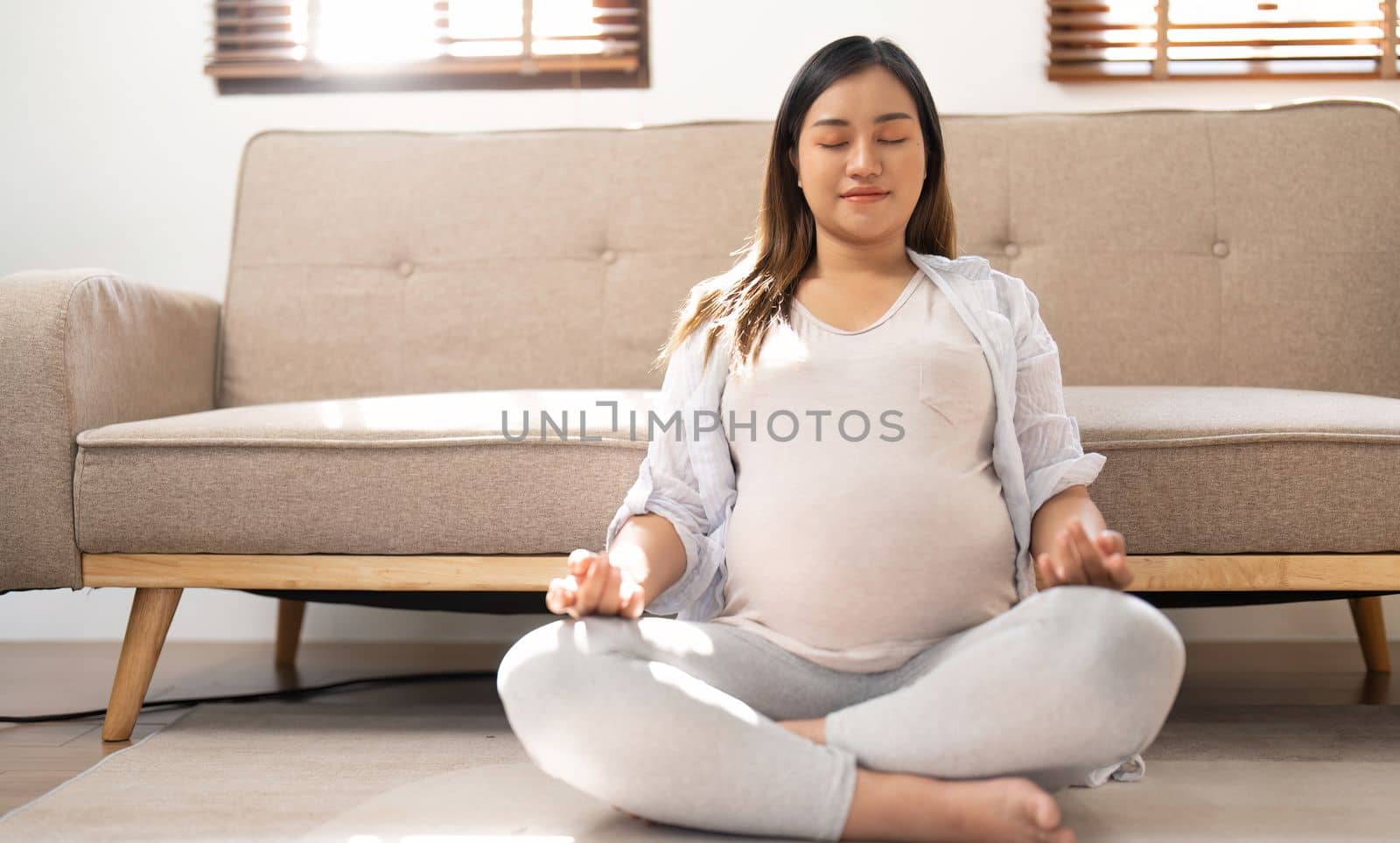 Relaxed Asian pregnant woman meditating in her living room, lotus pose, concentrating breath.