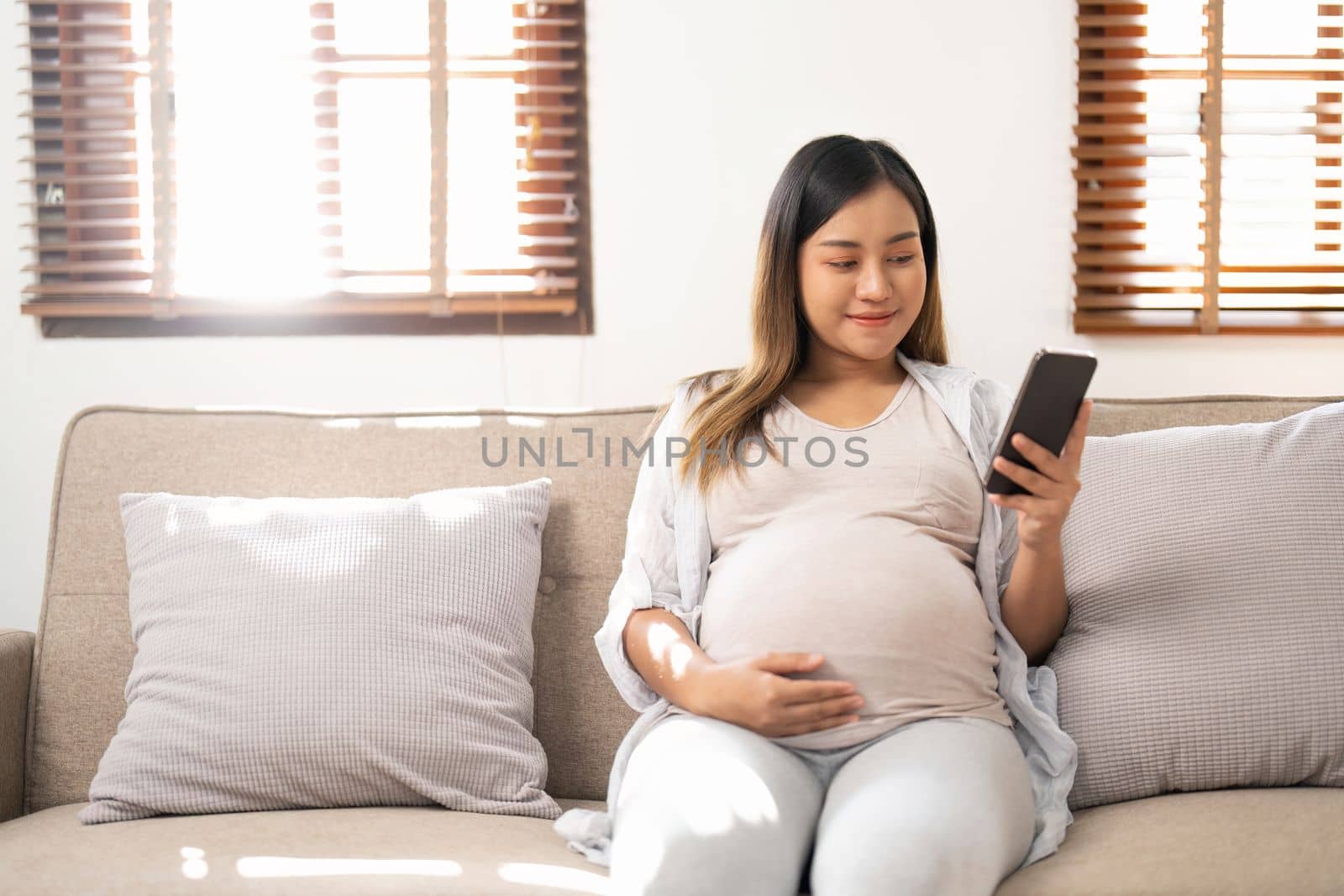 Happy Asian pregnant woman using her phone while relaxing on sofa in her living room by nateemee