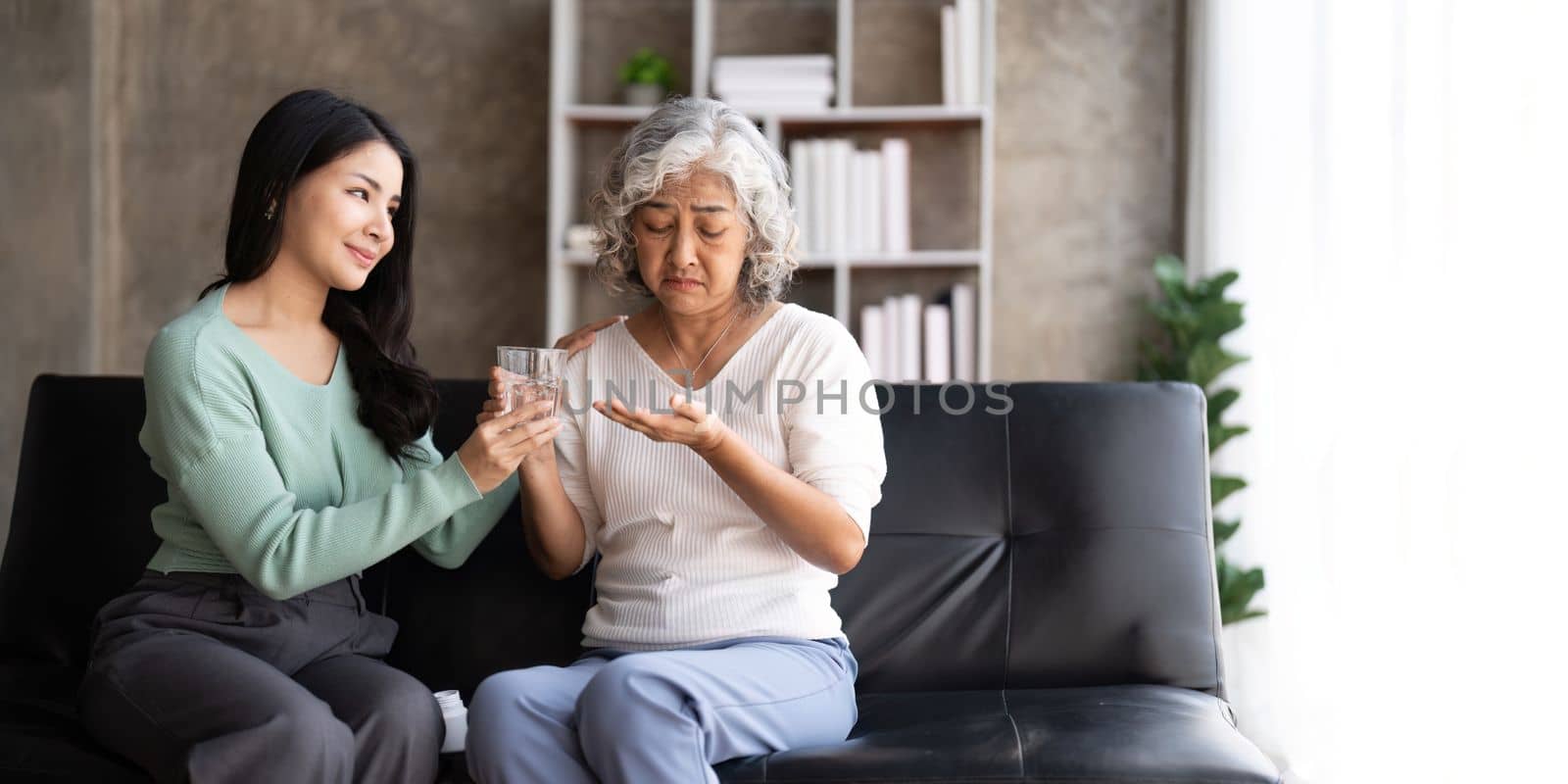 Asian daughter giving pills to mother helping her mom taking medicine with glass of water by nateemee