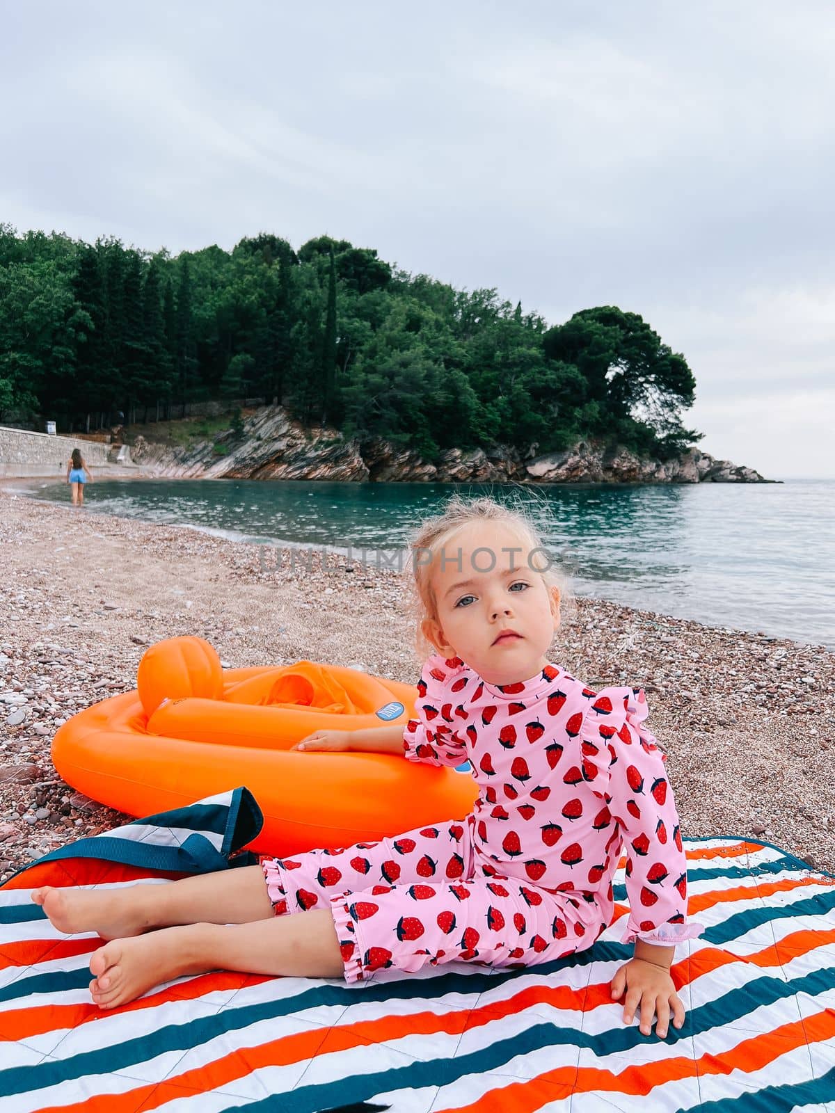 Little girl sits on a blanket on a pebble beach with her hand on an inflatable ring by Nadtochiy