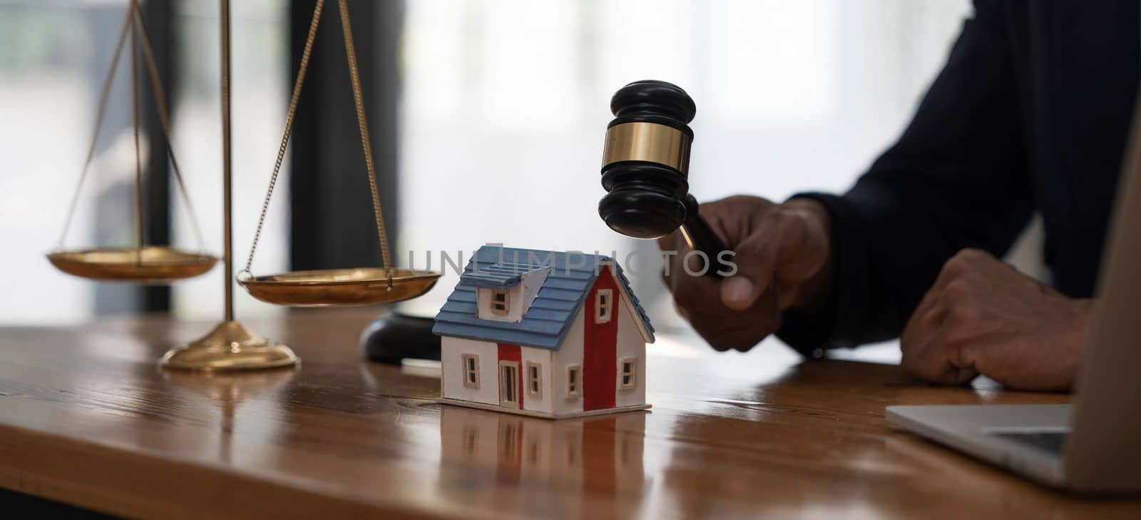 Close-up of a house in front of a lawyer holding a hammer and a laptop silver brass scales on a wooden table in his office, law, legal services, advice, justice and real estate ideas. by wichayada