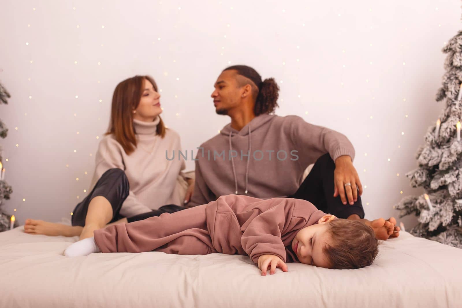 A little boy in a brown house suit , sleep on the bed, young parents kisses in the background, selective focus. Copy space