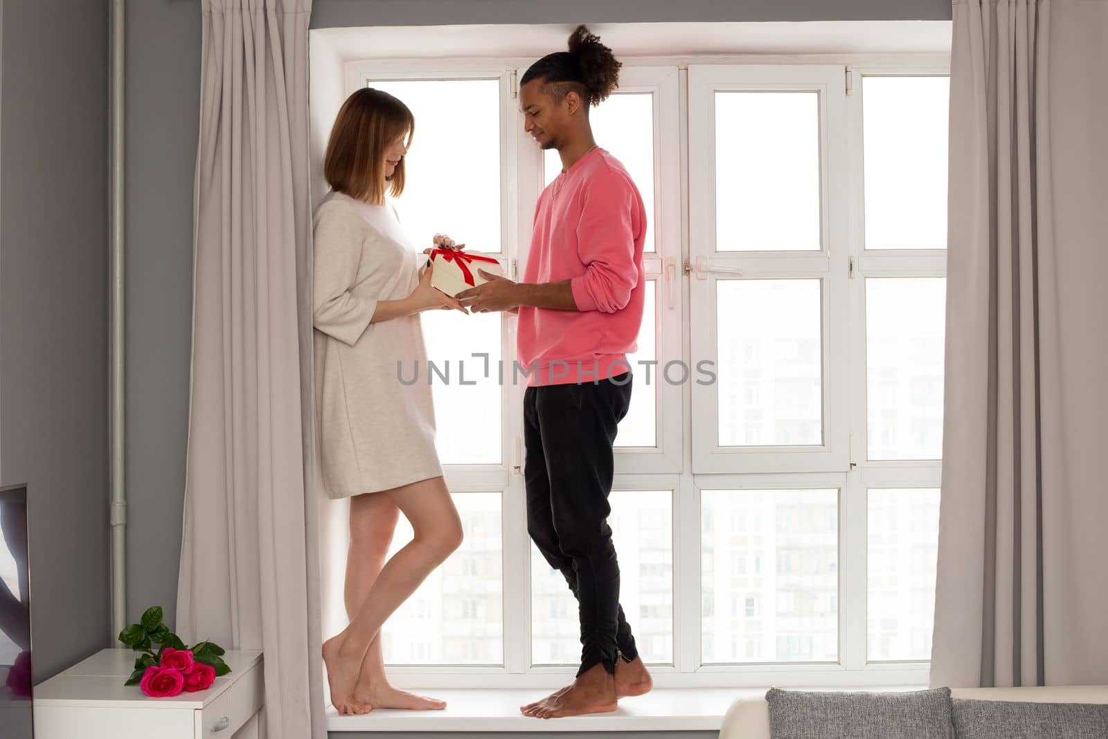 A guy in a pink jumper and a girl in a white dress are standing at a large white window in the room. by Zakharova