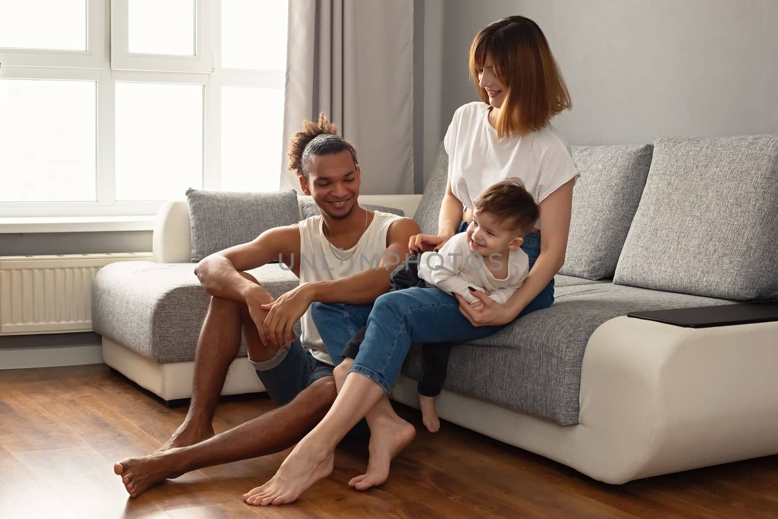 Happy, modern, multiracial family with a little boy in cozy home clothes sit on a gray sofa near a large window, laugh. Copy space