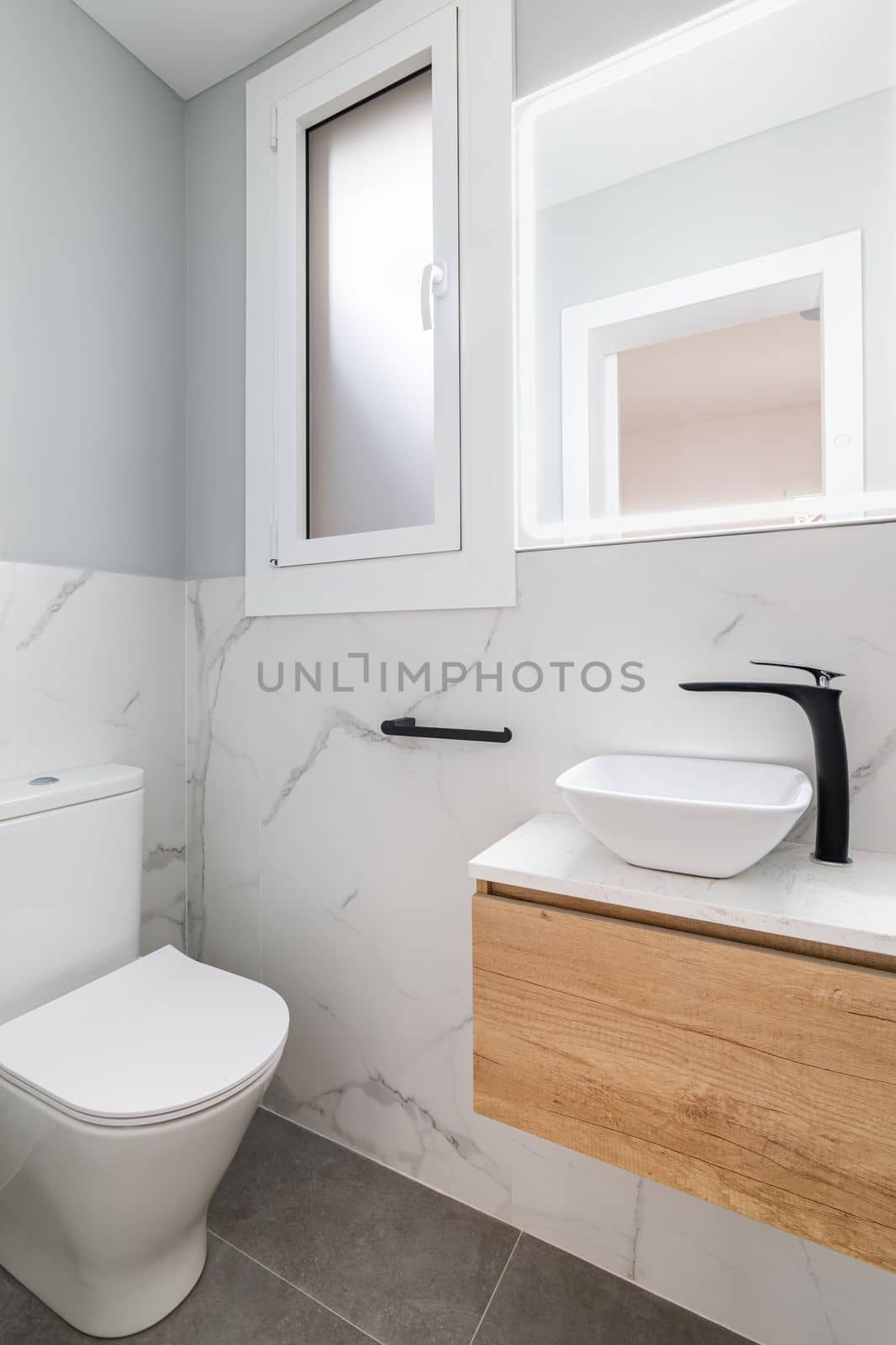Small bathroom with toilet and designer sink on small vanity with black faucet. Walls of room are made of white natural marble tiles. Square mirror is illuminated with fluorescent light