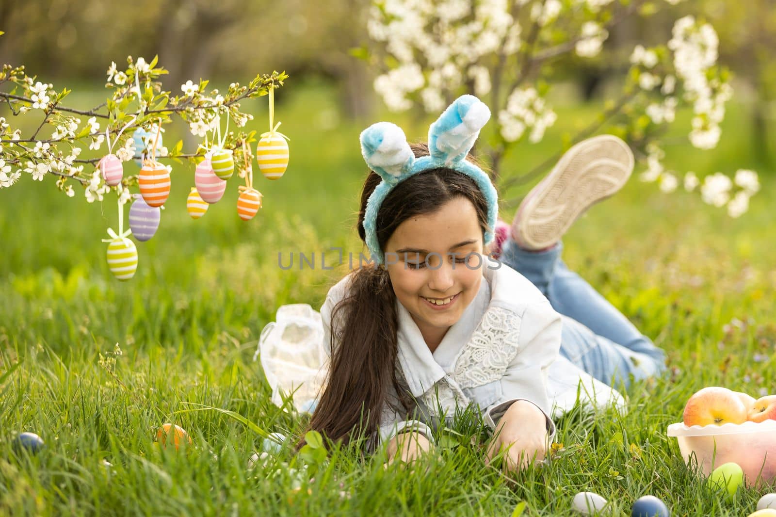 Adorable little girl in bunny ears, blooming tree branch outdoors on a spring day. Kid having fun on Easter egg hunt in the garden by Andelov13