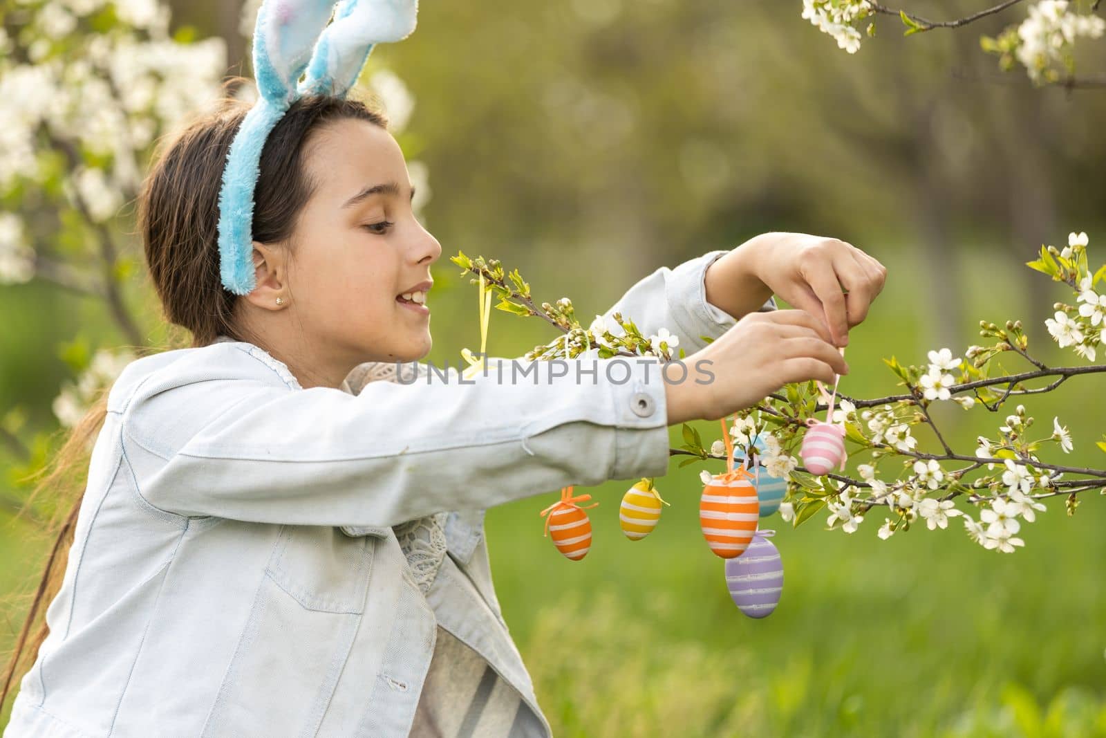 Adorable little girl in bunny ears, blooming tree branch outdoors on a spring day. Kid having fun on Easter egg hunt in the garden by Andelov13