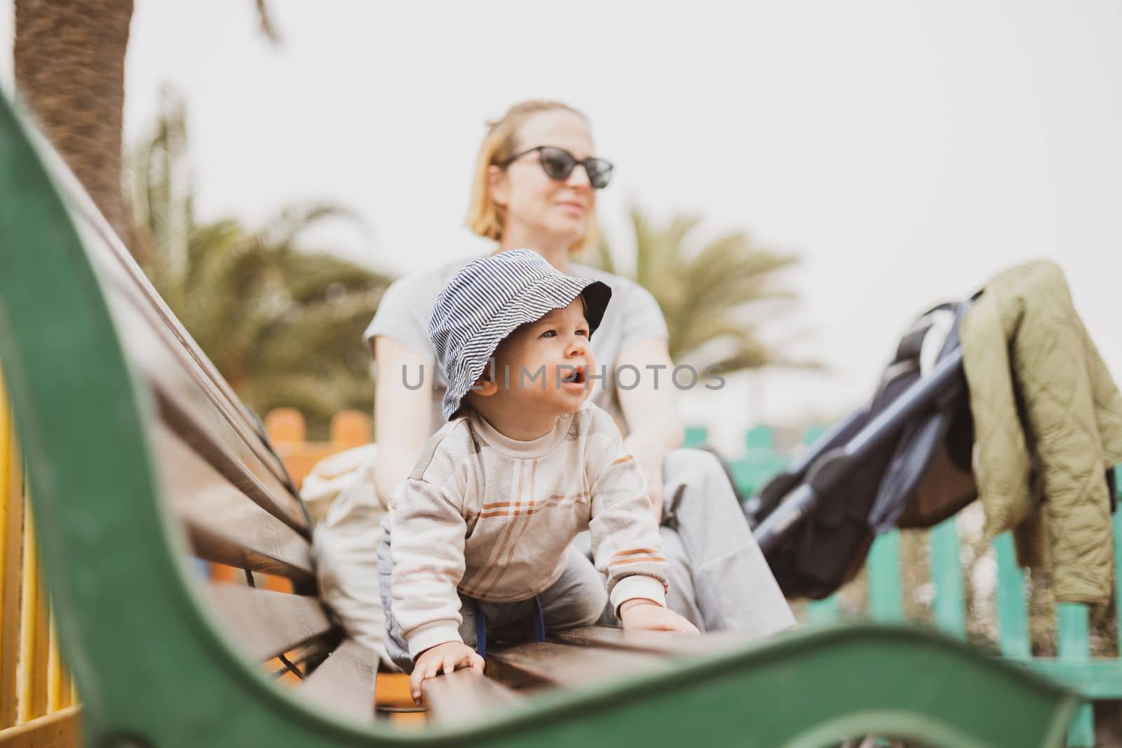 Young mother with her cute infant baby boy child on bench on urban children's playground on warm summer day
