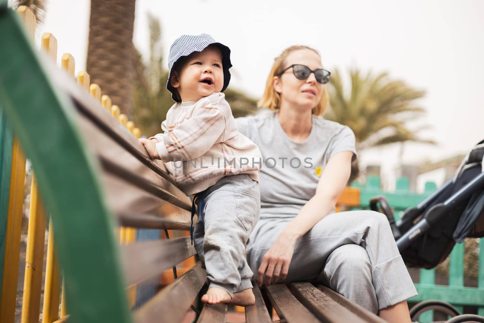 Young mother with her cute infant baby boy child on bench on urban children's playground on warm summer day