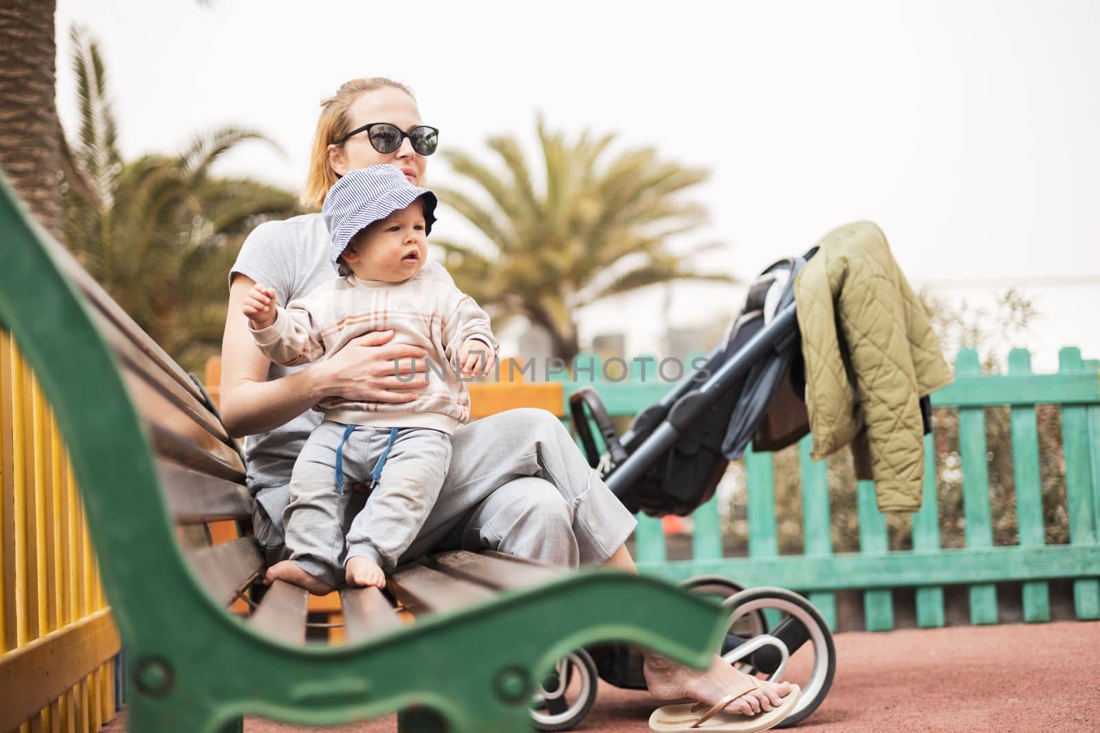 Young mother with her cute infant baby boy child on bench on urban children's playground on warm summer day