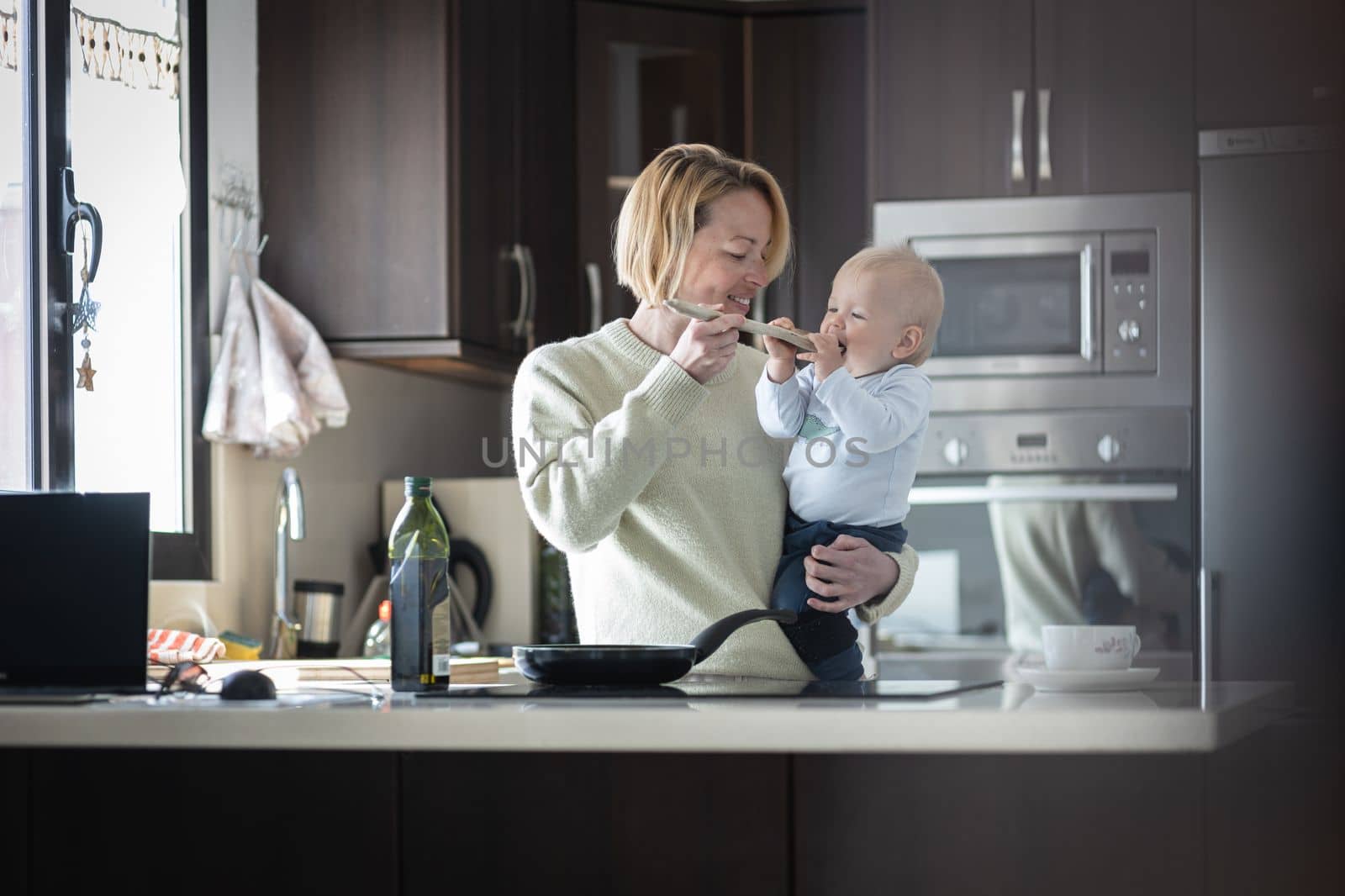 Happy mother and little infant baby boy cooking and tasting healthy dinner in domestic kitchen. Family, lifestyle, domestic life, food, healthy eating and people concept. by kasto