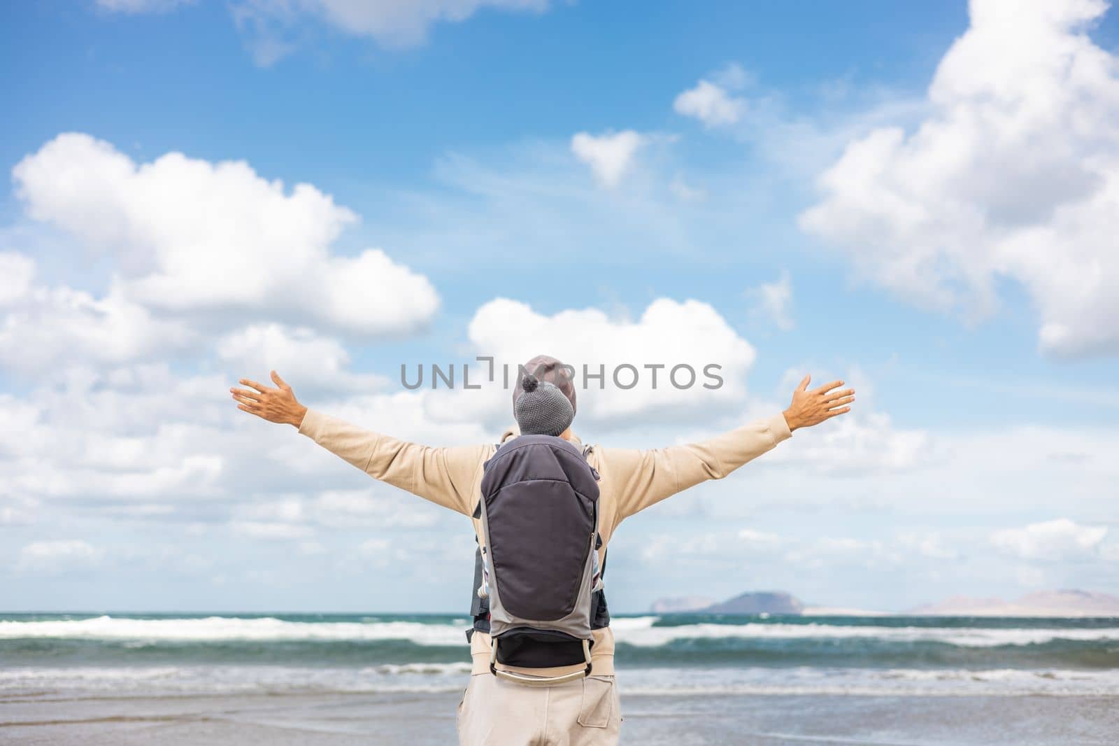 Young father rising hands to the sky while enjoying pure nature carrying his infant baby boy son in backpack on windy sandy beach of Famara, Lanzarote island, Spain. Family travel concept
